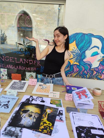 photo de moi (une femme blanche, longs cheveux noirs, en jean et débardeur noir) en train de prendre la pose derrière un stand présentant des illustrations, fanzines, linogravures que j'ai faites.