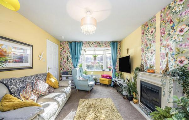 Screenshot of a property listing photo from Rightmove. A long narrow living room has been painted canary yellow. The end wall with a bay window has been wall papered on the flat outer wall with garish shiny large format floral wall paper, the bay is canary yellow. As if that weren’t bad enough the carpet is dirty brown and there are two random strips of the same garish wallpaper slapped on the wall either side of an enormously too-big-for-the-room faux mantlepiece which is itself festooned with plasctic pot plants. There appears to be a dead seagull nailed to the wall next to the light switch and to watch the TV you’d have to open the lounge door and sit in the hall… the furniture is smeared with ‘brown stains’ of dubious origin… 🤢