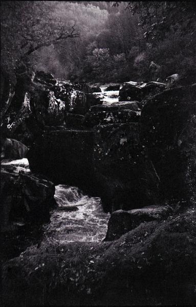Black and white photo of a boulder-bounded waterfall in a forest.  Overall the picture is dark apart from glimpses of white water and a slight shimmer of silver leaved trees to the top left.  A figure is barely distinguishable standing on the rocks at the top of the waterfall and fading into the forest.  Were they there and corporeal, or...?