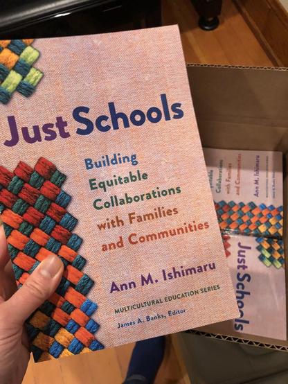 Hand holding Book titled Just Schools: Building Equit Collabs w/Families & Communities