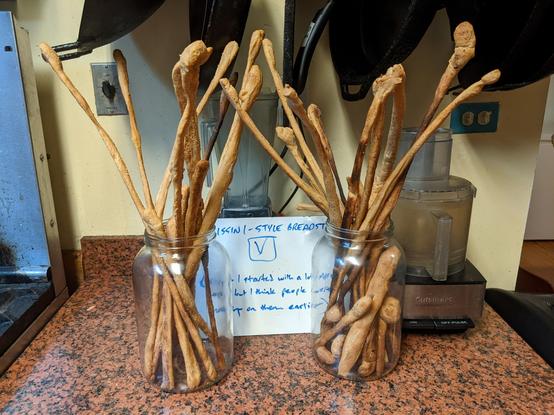 Two glass jars full of grissini-style thin Italian breadsticks.