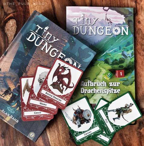 Das Bild zeigt einige Regelbücher und Spielkarten des Tiny Dungeon Systems, die auf einem Holztisch liegen. Zwei Sechserwürfel als Spieltools sind auch dabei.