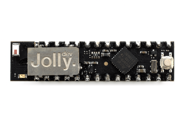 The Jolly Module, a small printed circuit board in a slightly elongated dual-inline pin (DIP) format, designed to replace the DIP-packaged ATmega328 chip which runs Arduino Uno and compatible microcontroller boards, seen from the top on a white background. A small metal shield bearing the Jolly logo is visible to the left.