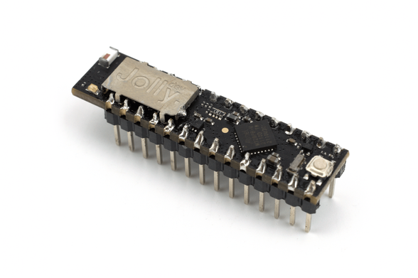 The Jolly Module, a small printed circuit board in a slightly elongated dual-inline pin (DIP) format, designed to replace the DIP-packaged ATmega328 chip which runs Arduino Uno and compatible microcontroller boards, seen at an angle on a white background. A small metal shield bearing the Jolly logo is visible to the left.