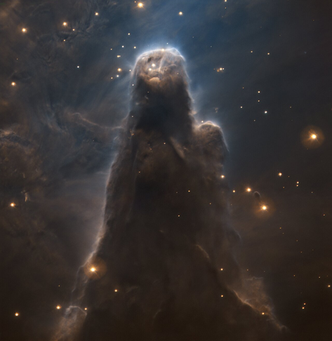 The image shows the Cone Nebula at the centre of the picture taking up most of the frame. The shape of the dark nebulous cloud is that of a cone, pointing upwards. At the top and surrounding the cone are bright yellow stars of different sizes, tiny dots and bigger gassy spheres. To the left of the central figure, abstract dusty patterns are fading upwards. The top shines brightly, lit up by luminous stars.