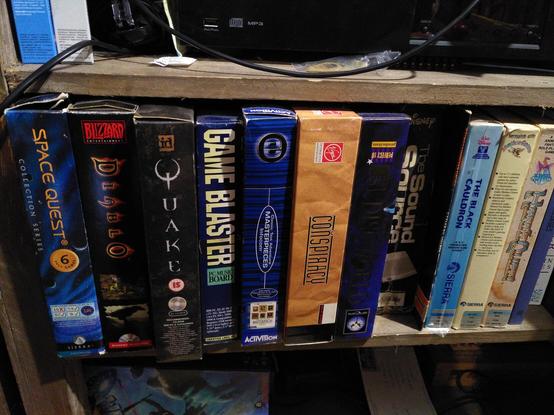 A row of boxes for games and hardware including Space Quest Collection, Diablo, Quake, a CMS Game Blaster, Infocom Masterpieces, Conspiracy, Discworld, a Covid Sound Source, a copy of Weird Computer Games, The Black Cauldron, Mixed Up Mother Goose, Hero's Quest, and a copy of Twisty Turny Passages