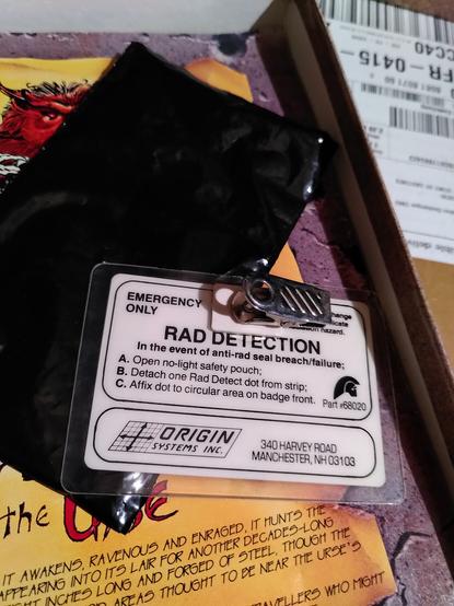 Rear of badge shows Origin systems logo and reads

RAD DETECTION
in the event of anti-rad seal breach/failure
A. Open no-light safety pouch
B. Detach one Rad Detect dot from strip
C. Affix dot to circular area on badge front