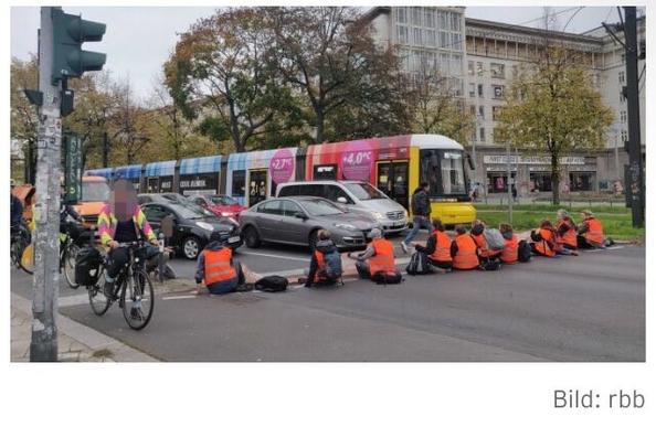10 Menschen in orangener Weste sitzen auf dem Fußgängerweg und blockieren Autos. Radfahrer fahren links vorbei, die Sraßenbahn rechts davon.