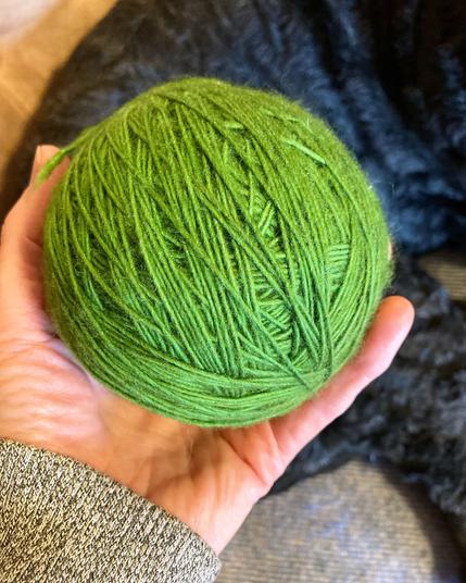 A hand holding a large ball of green yarn, the hind quarters of a black puppy in the background.