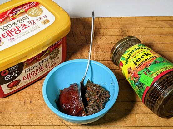 Picture of tub of Gochujang, bottle of Walkers Wood Jerk Sauce with a bowl in the middle. There's a 'dollop' of each in the bowl with a spoon forming a dividing line between them.
