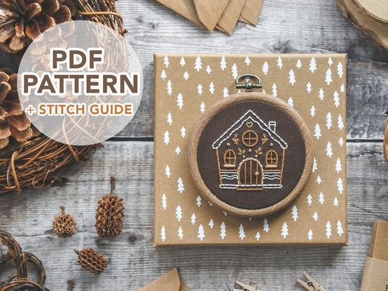 Photo of a hand embroidered Gingerbread House on brown fabric in a wood effect hoop frame. It’s placed on a light brown box with white trees on it. In the top left corner there is text saying ‘PDF Pattern and stitch guide’