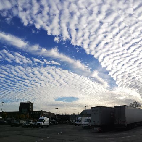 Schäfchenwolken, unterbrochen von einem freien Streifen blauen Himmels mit einem Kondensstreifen, der von links oben nach rechts unten führt, in weiterer Entfernung ein rundes Wolkenloch