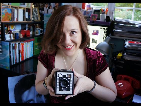 Jen sits in her messy office filled with memorabilia from film & tv, wearing a burgundy red velvet top, smiling at the camera and holding an vintage box brownie film camera from the 70s.