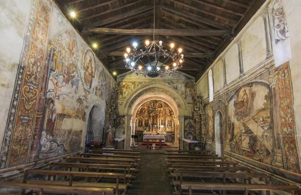 Interior de igreja pequena com trabes de madeira e paredes cobertas com mural colorido.