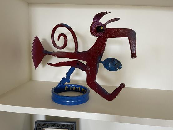 Folk art fish made with tool bits.  A vice grip body and mouth, fork tongs for eyebrows a wedge of circular saw for a tail and a spiral protruding from its back, all painted deep red except the chipped spoon tongue and stand which are painted blue