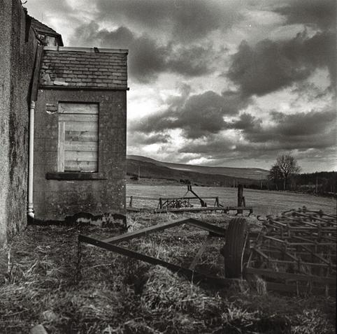 Black and white photo, overall muted and sullen without too much contrast.  In the left third can be seen a pebbledash wall of an abandoned building, a back-porch with a boarded up window makes up most of what is visible.  Where the porch meets the main wall there is a prominent white painted drainpipe.  In the bottom right there is some old abandoned and rusting farm machinery (think it's a scarifier plough).  Above that a sliver of lighter field, then a darker sliver of hills in the distance.  The sky above has dark clouds with bright edges and looks stormy.