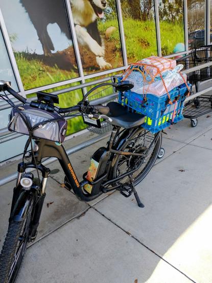 A black ebike with a blue plastic milk crate on the rear rack. The crate is stuffed full of shopping bags and strapped down with bungee cords.
