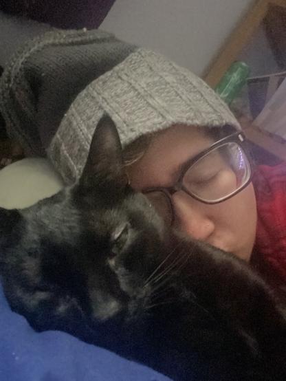 Small black cat with green eyes partially open, accepting cuddles from his human who is feeling UNWELL
