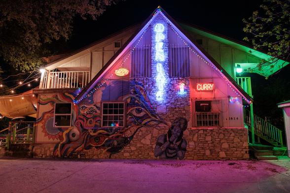 A night-time photo of a building with an abstract Indian mural showing Shiva smoking a chillum with birds for smoke. The building also has many neon signs on it, with the most prominent one saying "open late".