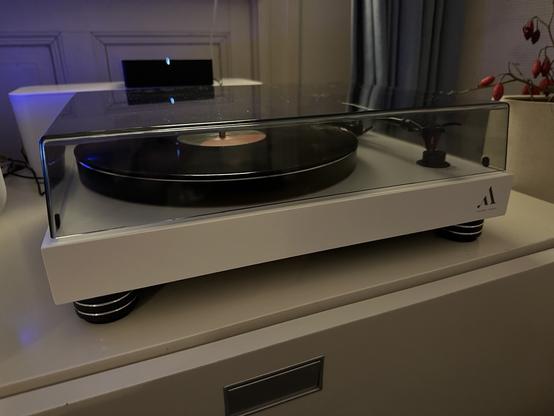 Analog record player in white with a record on the turntable. The transparent hood is closed.