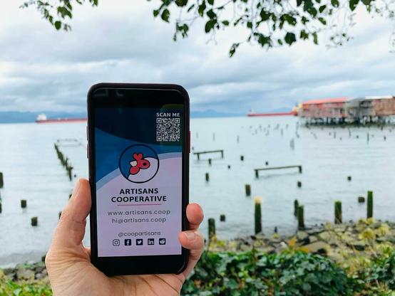 A phone being held in front of the Colombia River displaying an Artisans Cooperative digital business card.