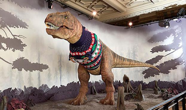 Picture of the animatronic t-rex model at the National History Muesume in London wearing a dark blue Christmas sweater that has stripes with different designs that go all the way around the sweater: one is of white snow against the dark blue, white snowflakes against a red stripe, green dinosaurs, a red stripe with white snow, green christmas trees against a white stripe, then the pattern repeats again.