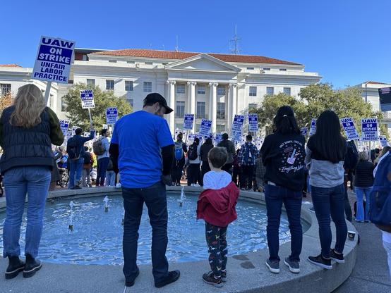 Picture of people standing on Ludwig’s Fountain with a crowd looking at Sproul Hall at UC Berkeley. Picket signs with “UAW ON STRIKE UNFAIR LABOR PRACTICE” held high.