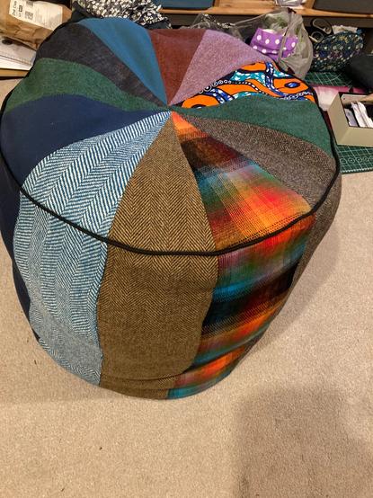 A range of triangular fabric pieces are joined to create a circular patchwork top to a 60cm tall cylindrical pouf. The fabrics are a mix of wool and brushed cotton and African wax print. Taken from a different angle to photo one to show off the different range of fabrics and colours.