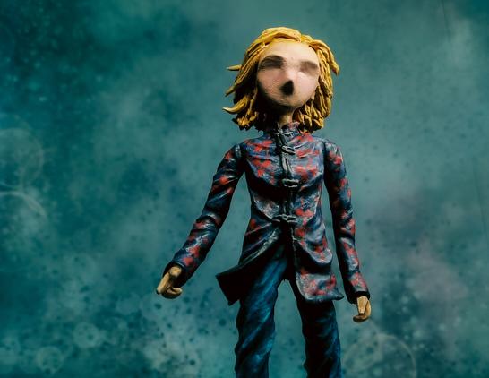 Handmade clay figurine, tall and skinny, stylised with minimal facial features. Based on the character of the 13th Doctor Who. Wearing a dark navy coat with red flowers over her short blue trousers. The background is dark green and feels like it's deep under the sea.