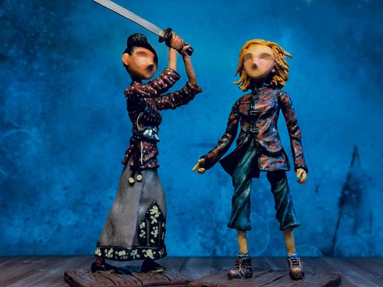 Two handmade clay figurines, one of Yaz and one of the 13th Doctor. Stylised, tall, skinny, minimal facial features. Image is a full body view. Dark blue background.