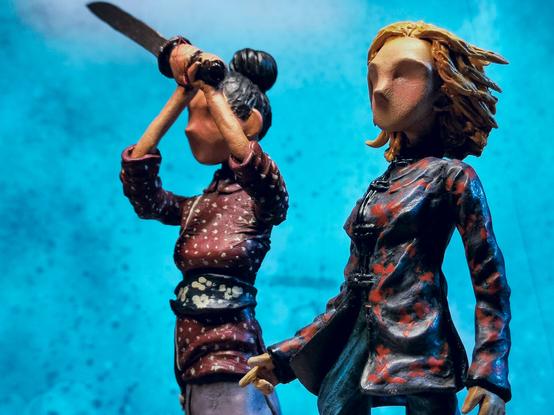 Two handmade clay figurines, one of Yaz and one of the 13th Doctor. Stylised, tall, skinny, minimal facial features. Image is a half body view. Bright blue background.