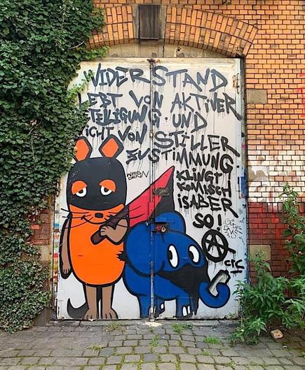 Auf dem Bild ist ein Graffiti mit einer vermummten Maus und dem Elefanten, der beliebten Kindersendung, zu sehen. Der Elefant hat eine Graffitidose im Rüssel und die Maus eine Schwarz/Rote-Fahne in der Hand. Hinter beiden steht ein Text: Widerstand lebt von aktiver Beteiligung und nicht von stiller Zustimmung. Klingt komisch ist aber so!