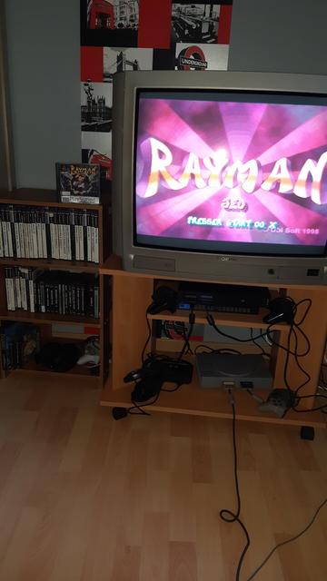 Photo of my game corner, with a big crt monitor displaying the totle screen of Rayman on the playstation 1. under it, a ps2, ps1 and sega genesis can be seen. to the left of it, a small shelf with ps2 and ps1 games as well as a few controllers.