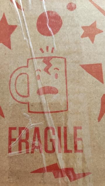 Eine gezeichnete Abbildung einer Tasse, mit einem Gesicht. Das Gesicht reißt die Augen auf und schaut geschockt, ob eines Risses an ihrer Oberkante.
Darunter steht "Fragile".
Un die Tasse herum sind Sterne, Kreise, Dreicke und Blitze abgebildet, ebenfalls in Rot.