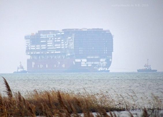 Der Rohbau des Mittelschiffs eines Kreuzfahrers bei diesigem Novemberwetter auf dem Wasser der Ostsee, umgeben von mehreren Schleppern. Küstendünen und braunes Gras im Vordergrund.