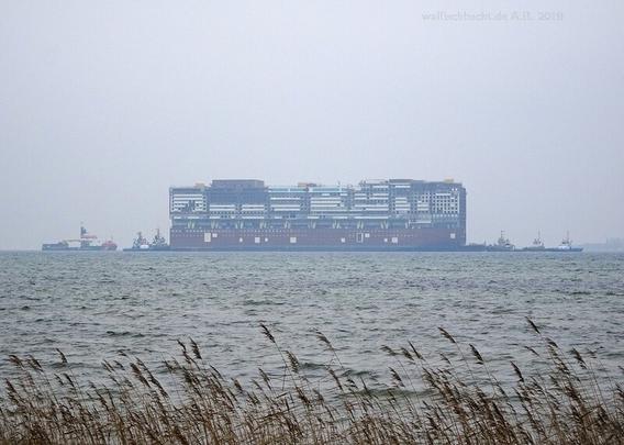 Der Rohbau des Mittelschiffs eines Cruiseliners bei diesigem Novemberwetter auf dem Wasser der Ostsee, umgeben von mehreren Schleppern und Sicherungsfahrzeugen.