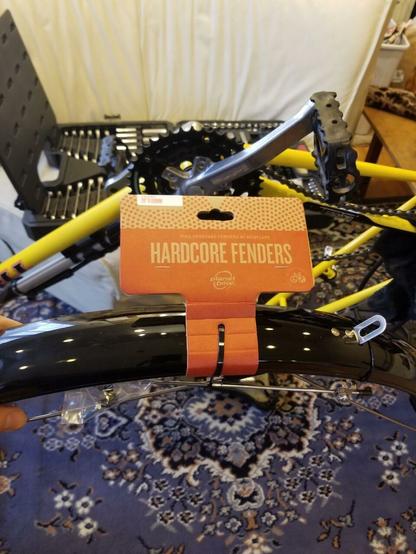 An unopened package of Planet Bike Hardcore Fenders is visible in front of the frame of a yellow bicycle (with wheels removed) and a socket/wrench set in the background.