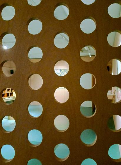 Round holes in a wood panel with the exhibition space behind, and the screened picture in the background.