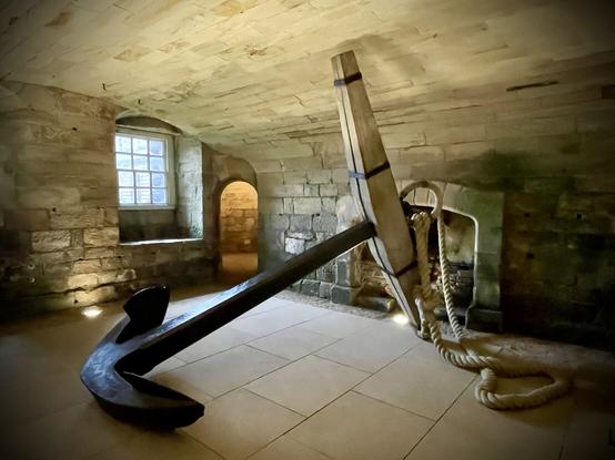 A low-ceilinged room with stone walls, floor and arched roof. There is a stone fireplace at one end and an arched doorway in the far corner through which artificial light spills. In the middle of the floor, occupying most of the room and almost touching the ceiling is a giant wonder anchor with a hefty rope through a ring at the top.