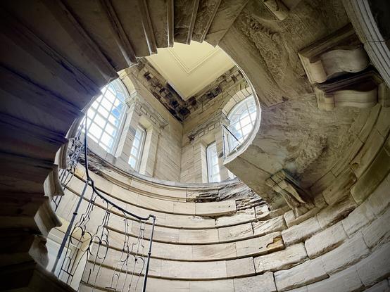 A broken stone spiral staircase in a grand scale, shown from below, with bent iron railings. It ends abruptly, but above it are large arched windows and carvings at the roof edge.