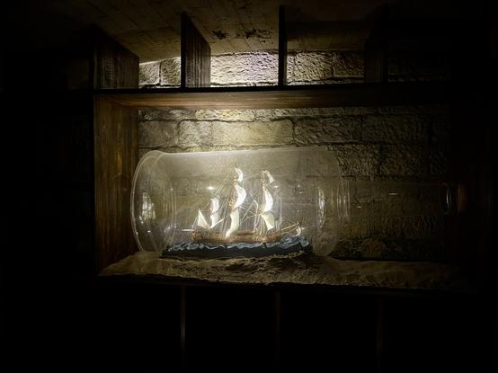 A dark room with a wooden cabinet against a stone wall. Resting on a shelf in the cabinet is a giant illuminated glass bottle, around three to four feet long, containing a sailing ship with white unfurled sails.