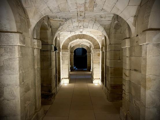 A long stone corridor with arched doorways leading off to the left and right. At the end of the corridor, through another arch, is a flight of stone steps leading up to the left and bathed in dark blue light.