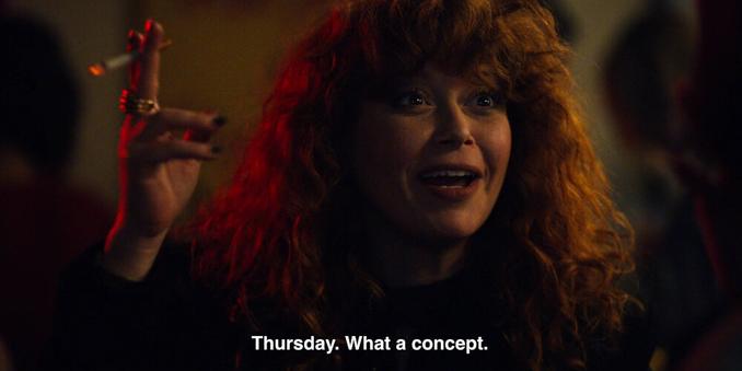 A screenshot from the series Russan Doll. Woman with long brown wavy hair is holding a cigarette and is talking to someone in front of her. Captions read: "Thursday. What a concept.