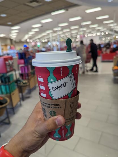 Paper Starbucks holiday cup with my name (August) written on it. In the background, blurry, is the Target store.