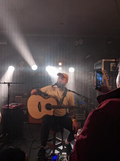 A photo of stage mid-performance. A man in his 20's, dressed in a tee-shirt and baseball cap, sings into a microphone and holds an acoustic-electric guitar.
