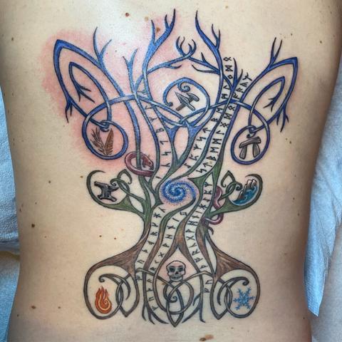 Photo of a large back tattoo in the shape of a knotwork tree which shades from brown at the roots, to green at the center, to blue in the branches. Rows of runes snake along its length and icons of the nine Norse worlds are held among its roots and branches.
