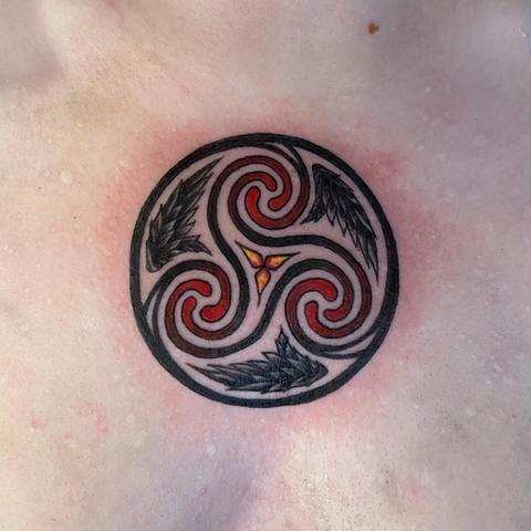 Photo of a triskelion tattoo, with a triple spiral in red shading to black at the outer edges, joined to a black outer ring. Small black wings decorate the spaces between the three spirals.