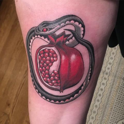 Photo of a tattoo which centers a pomegranate split open showing the shiny red seeds. A snake coils around it with a gray and black patterned body and white underbelly.