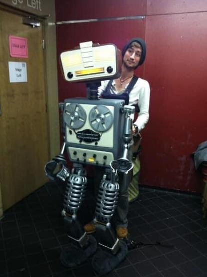 Photo of the Marvin puppet held up by his operator standing behind. The puppet is very Heath Robinson style, with separate head and body, and arms and legs that look as though they are repurposed pieces of equipment. The torso includes a reel to reel tape machine. Very retro feel.