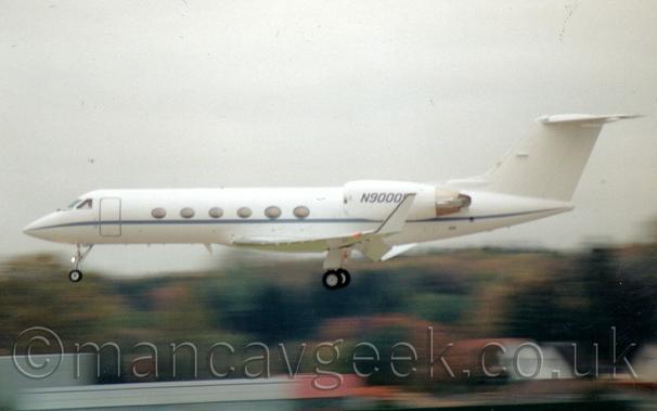White bizjet shortly before landing at an airport.
In tha background are some trees and a building, under a grey sky.